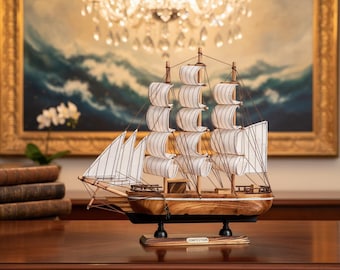 Op de afbeelding: Gedetailleerd houten model van een zeilschip met witte zeilen en een bruine houten romp. Het schip staat op een houten voet met het woord "CONFECTION". De achtergrond omvat een schilderij en een kroonluchter.