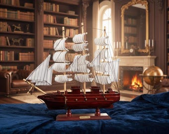 Op de afbeelding: Gedetailleerd houten model van een schip met witte zeilen en een donkerrode romp. Het schip staat op een blauw oppervlak, met een bibliotheek en een open haard op de achtergrond. Het model is een decoratief nautisch item.