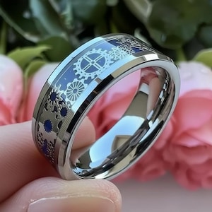 May include: A silver ring featuring a blue and silver gear design. The ring has a polished, reflective surface and a blue inner band with gear and cogwheel patterns. The ring is held in a hand, with pink flowers in the background.