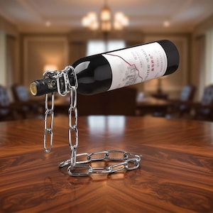 Puede incluir: Una botella de vino suspendida en un soporte de cadena plateada. La botella es oscura con una etiqueta blanca. La cadena está conectada a la botella y descansa sobre una mesa de madera oscura. El fondo es una habitación borrosa.