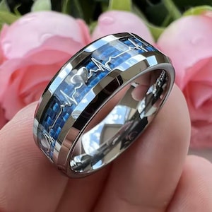 Puede incluir: Un anillo plateado con una incrustación de fibra de carbono azul y un diseño de latido cardíaco. El anillo tiene un acabado pulido y está sobre un fondo de flores rosas. El diseño del anillo presenta un patrón de latido cardíaco.