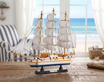 Op de afbeelding: Een decoratief model van het schip "Cutty Sark", met witte zeilen en een witte romp met gele en blauwe accenten. Het schip staat op een houten tafel, met een strandscène op de achtergrond.