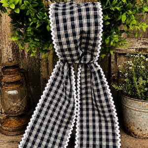 May include: A long fabric ribbon featuring a navy blue and cream plaid pattern with white rickrack trim along the edges. The ribbon is draped vertically, showcasing the woven textile texture and the consistent wavy white border detail.