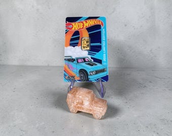 May include: A rectangular Hot Wheels 2026 Limited Edition card featuring a light blue pickup truck graphic and a small gold bar embedded in the center, positioned above a translucent, light peach-colored carved stone vehicle figurine.