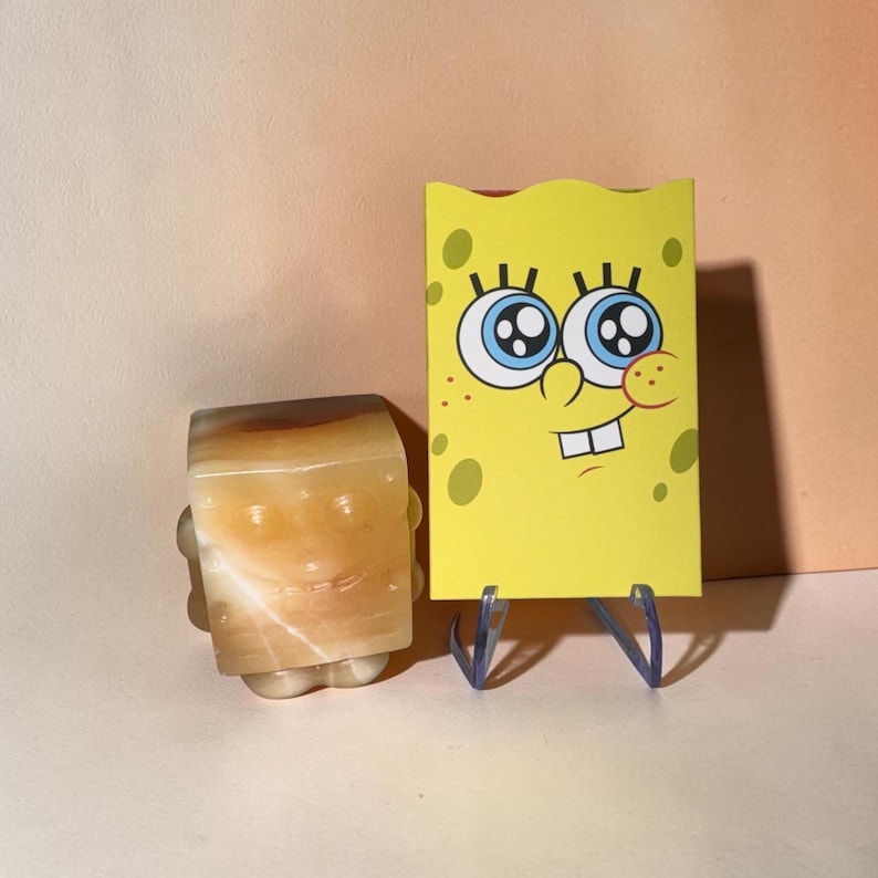 May include: A yellow SpongeBob SquarePants card with blue eyes and a smiling face stands next to a carved stone figure also resembling SpongeBob. The stone figure is beige and brown with a smiling face.