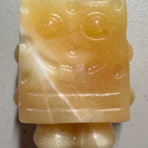 May include: A carved, yellow and white stone figurine of a cartoon character with a square body, large eyes, and a smiling mouth. The figurine has a small, rounded base and is made of a translucent material.