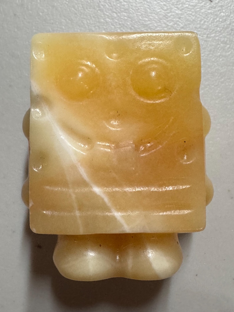 May include: A carved, yellow and white stone figurine of a cartoon character with a square body, large eyes, and a smiling mouth. The figurine has a small, rounded base and is made of a translucent material.