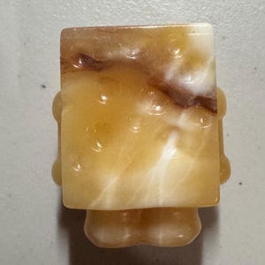 May include: A carved, abstract figurine in shades of yellow, cream, and brown. The figure has a rectangular head and a rounded body with small, rounded protrusions on the sides. The stone has a marbled appearance.