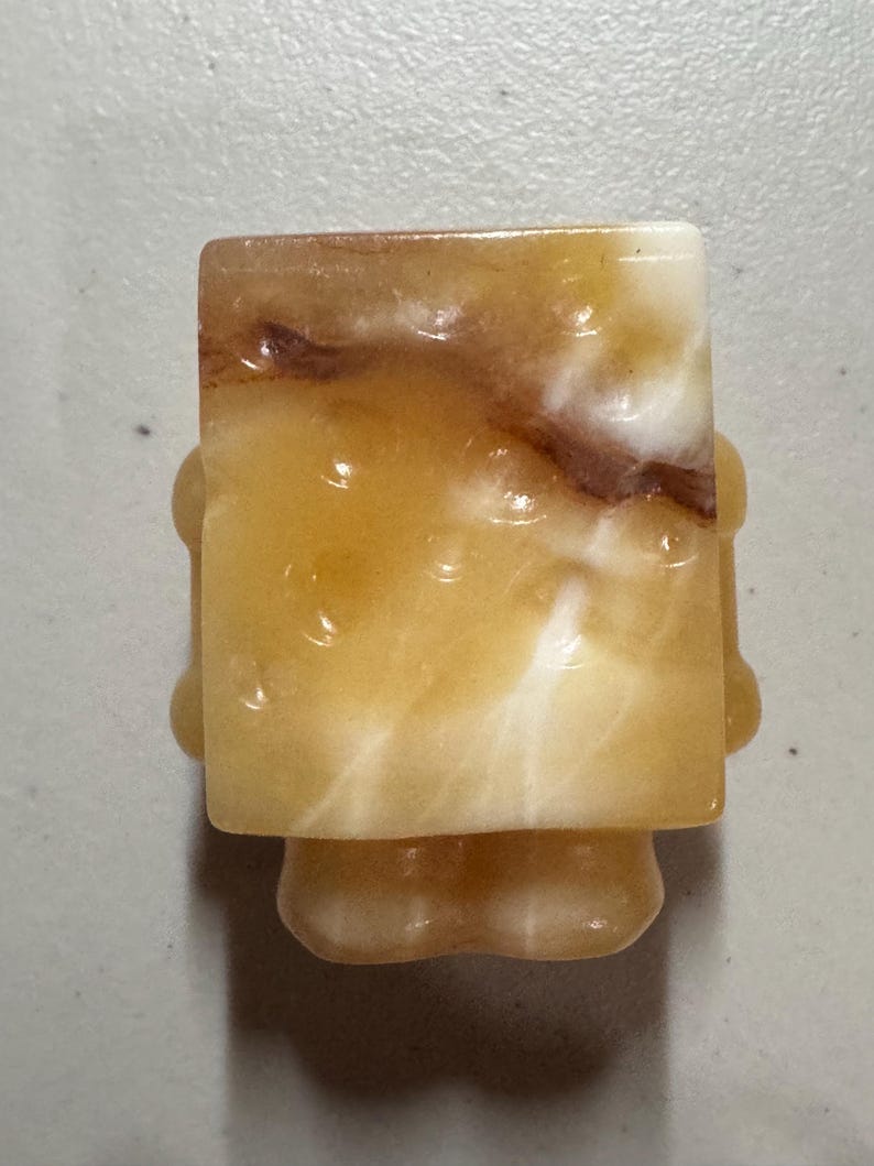 May include: A carved, abstract figurine in shades of yellow, cream, and brown. The figure has a rectangular head and a rounded body with small, rounded protrusions on the sides. The stone has a marbled appearance.