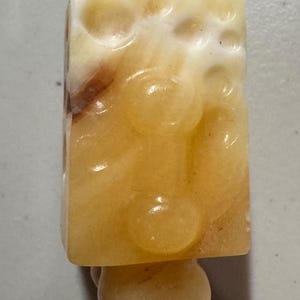 May include: A rectangular, light-yellow stone carving with a rounded base. The stone has a textured surface with circular indentations and brown veining. The base is a smooth, rounded shape.