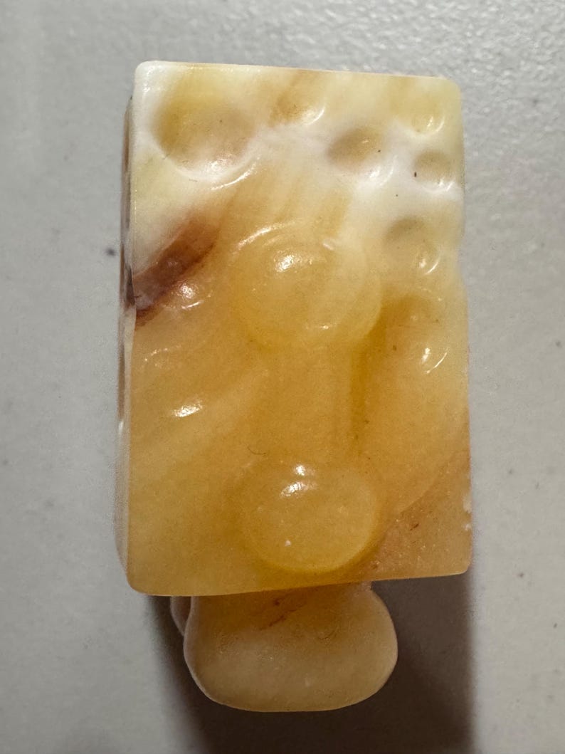 May include: A rectangular, light-yellow stone carving with a rounded base. The stone has a textured surface with circular indentations and brown veining. The base is a smooth, rounded shape.