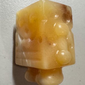 May include: A small, yellow-orange, carved stone figurine with a cubic head and rounded body. The stone has a translucent quality with darker brown inclusions. The figurine has a unique, abstract design.