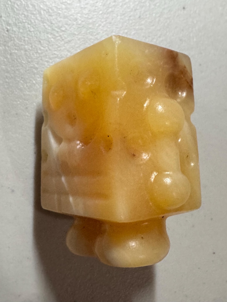 May include: A small, yellow-orange, carved stone figurine with a cubic head and rounded body. The stone has a translucent quality with darker brown inclusions. The figurine has a unique, abstract design.