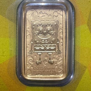 May include: A gold-colored rectangular bar featuring an embossed image of the cartoon character SpongeBob SquarePants. The bar is encased in a clear, rectangular protective case. The text "&copy; 2025 Viacom International Inc." is visible.