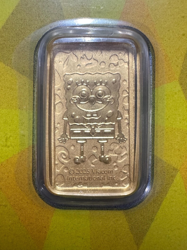 May include: A gold-colored rectangular bar featuring an embossed image of the cartoon character SpongeBob SquarePants. The bar is encased in a clear, rectangular protective case. The text "&copy; 2025 Viacom International Inc." is visible.