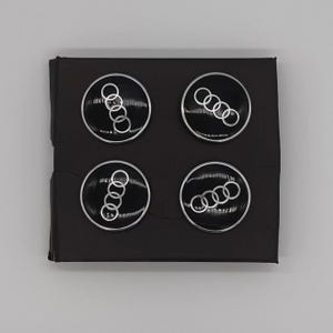 May include: Four black and silver car emblems arranged in a square. Each emblem features a silver ring design on a black background, set against a black backdrop.