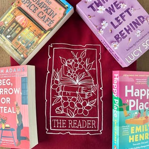 Op de afbeelding: Een bordeauxrode sweatshirt met een wit bloemen- en boekontwerp met de tekst "THE READER" is omringd door vier boeken. De boeken hebben kleurrijke covers en titels als "The Pumpkin Spice Cafe" en "Happy Place".