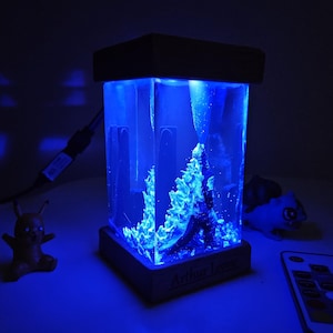 Könnte beinhalten: Ein rechteckiges, blau beleuchtetes Kunstwerk aus Kunstharz. Es zeigt eine detaillierte Unterwasserszene mit Korallen und einer dunklen Figur. Der Sockel ist aus Holz mit dem Namen "Arthur Lome". Eine Fernbedienung und Figuren sind in der Nähe.