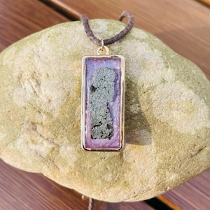 May include: A rectangular pendant necklace with a gold-colored frame. The pendant features a dark gray, textured design set against a purple and pink background. The necklace has a brown cord.