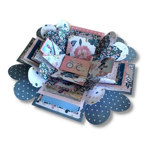 Peut inclure: Une boîte explosion artisanale à plusieurs couches avec des motifs floraux et à pois. La boîte présente des couleurs roses, bleues et blanches, avec des images de vélos et de fleurs. La boîte est ouverte, révélant plusieurs couches et compartiments.