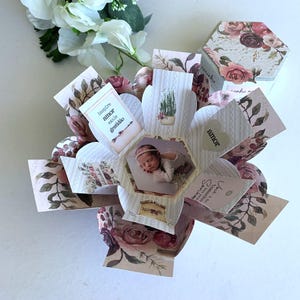 Puede incluir: Una caja explosiva hecha a mano con una foto de bebé en el centro. La caja está decorada con paneles florales y en forma de corazón, con las palabras "inspire amor exale gratidão" y "amor". Un regalo único.