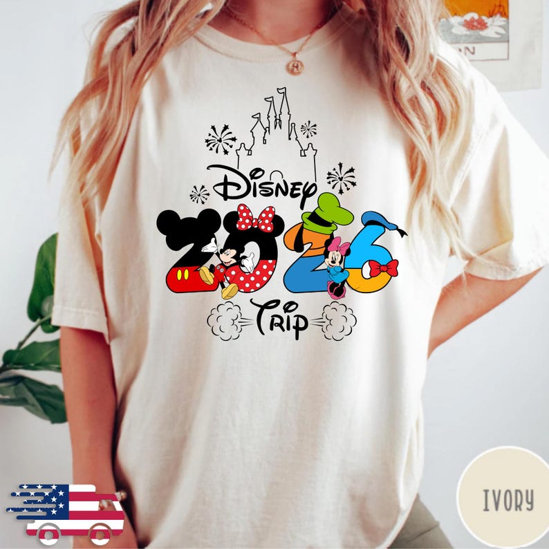 May include: An ivory t-shirt featuring a graphic design with the text Disney 2026 Trip. The numbers are stylised with Mickey Mouse, Minnie Mouse, Goofy, and Donald Duck motifs, topped with a black outline of a castle and firework accents.