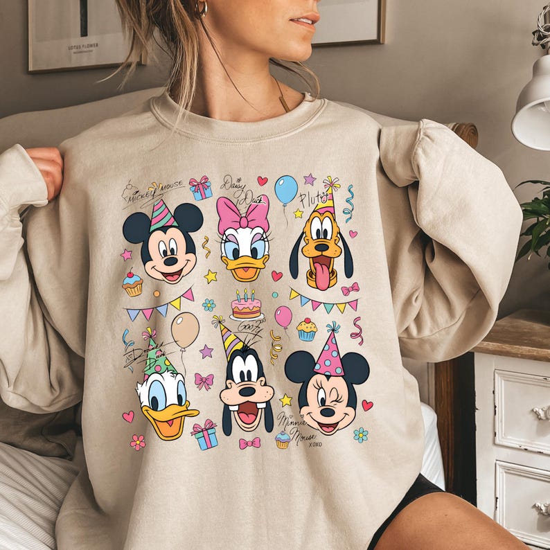 May include: A beige crewneck sweatshirt featuring a colourful graphic print of Disney characters including Mickey Mouse, Minnie Mouse, Donald Duck, Daisy Duck, Goofy, and Pluto wearing party hats, surrounded by birthday cakes, balloons, and gifts.