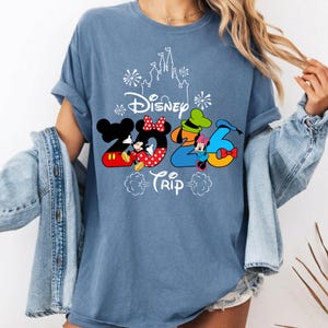 May include: A blue short-sleeve t-shirt featuring a white outline of a castle and fireworks above the text Disney 2026 Trip. The graphic includes Mickey Mouse, Minnie Mouse, Goofy, and Donald Duck integrated into the numbers 2026 in bright colours.