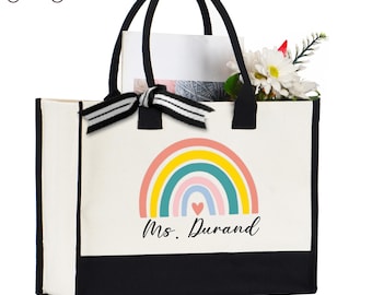 May include: A white canvas tote bag with a black bottom and handles. The bag features a colorful rainbow design with the text "Ms. Durand" and a heart. The bag is filled with white flowers and a bow.