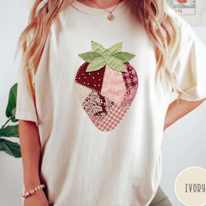 May include: Ivory-colored t-shirt featuring a patchwork strawberry design. The strawberry is composed of various fabric patterns in shades of red, pink, and white, with green leaves at the top. The shirt has a relaxed fit.