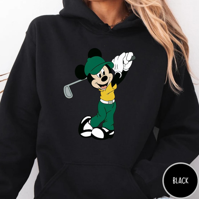 May include: A black hooded sweatshirt featuring a graphic print of Mickey Mouse dressed in a green cap, yellow shirt, and green pants, holding a golf club. The character is depicted in a golf swing pose with white gloves and black and white shoes.