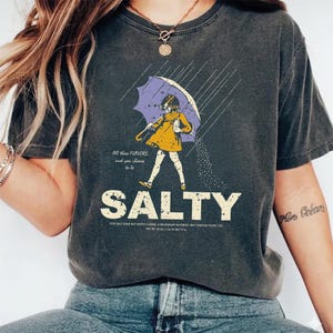 May include: Dark gray t-shirt with a graphic of a person under a purple umbrella, with the word "SALTY" in large white letters. The text "All these flavors and you choose to be" is above the word "SALTY".
