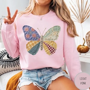 May include: Light pink sweatshirt featuring a large butterfly design. The butterfly has patchwork wings in various colors and patterns, including floral, plaid, and solid fabrics. The sweatshirt has long sleeves and a crew neck.