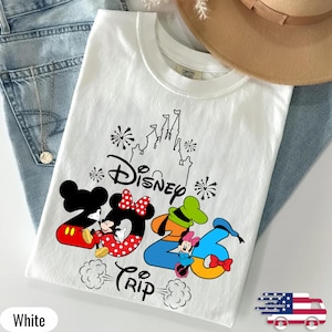 May include: A white crew neck t-shirt featuring a black graphic of a castle, fireworks, and the text Disney 2026 Trip. The design includes Mickey Mouse, Minnie Mouse, and Goofy integrated into the numbers, with red and blue colour accents.