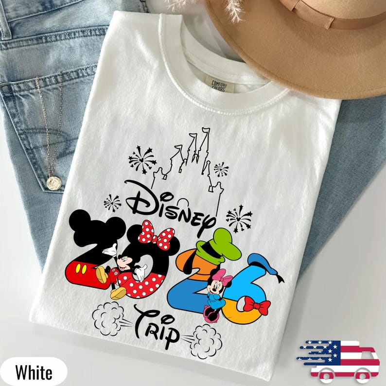 May include: A white crew neck t-shirt featuring a black graphic of a castle, fireworks, and the text Disney 2026 Trip. The design includes Mickey Mouse, Minnie Mouse, and Goofy integrated into the numbers, with red and blue colour accents.