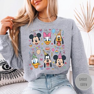May include: A heather grey crewneck sweatshirt featuring a colourful graphic print of Disney characters including Mickey Mouse, Minnie Mouse, Donald Duck, Daisy Duck, Goofy, and Pluto wearing party hats, surrounded by balloons, cupcakes, and gifts.