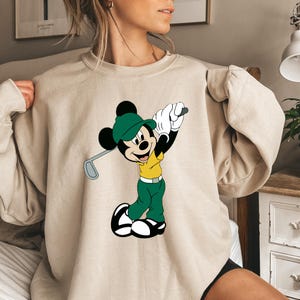 May include: A beige crewneck sweatshirt featuring a graphic print of Mickey Mouse wearing a green cap, a yellow polo shirt, and green trousers while holding a golf club. The character is depicted in a golf swing pose with white gloves and shoes.