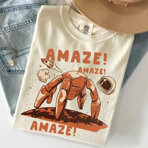 May include: Cream-colored t-shirt with an orange graphic of a crab-like creature on a planet, a rocket, and an astronaut helmet. The word "AMAZE!" is printed above and below the graphic. The shirt is folded on top of blue jeans.