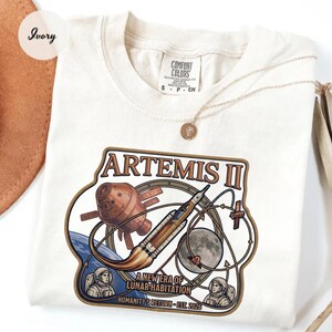 May include: Ivory-colored t-shirt featuring a graphic of the Artemis II mission. The design includes a rocket, spacecraft, Earth, and the moon, with the text "ARTEMIS II" and "A NEW ERA OF LUNAR HABITATION". The shirt is a Comfort Colors brand.