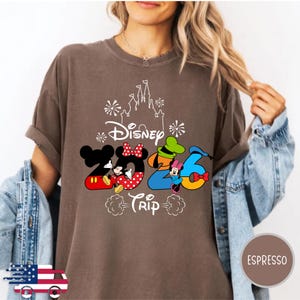 May include: A brown short-sleeve t-shirt featuring a white outline of a castle and fireworks above the text Disney 2026 Trip. The numbers 2026 are stylised with Mickey Mouse, Minnie Mouse, Goofy, and Donald Duck character elements in red, yellow, blue, and green.