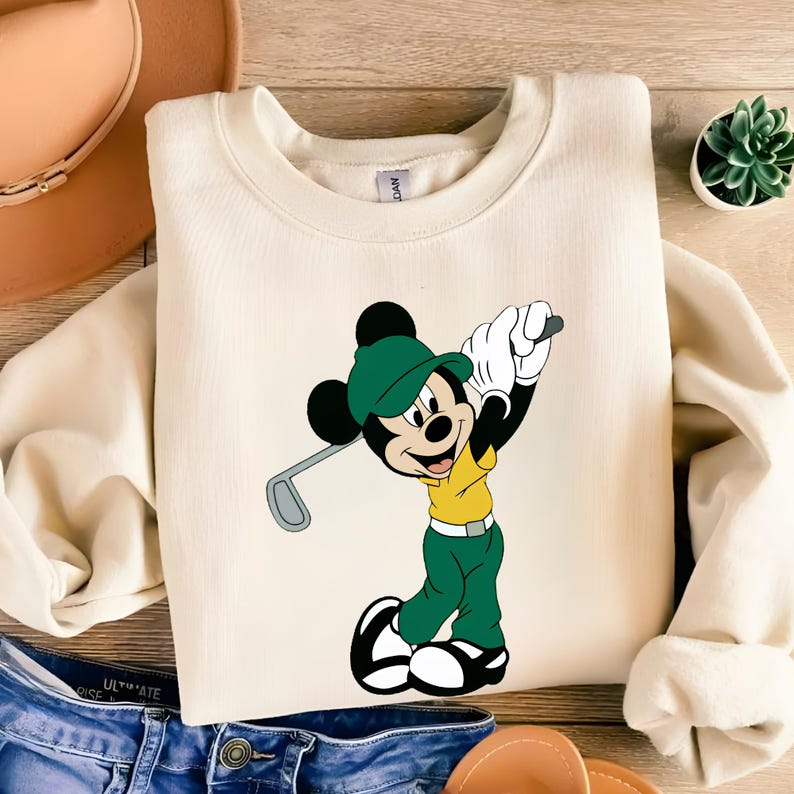 May include: A cream-colored crewneck sweatshirt featuring a graphic print of Mickey Mouse wearing a green golf cap, a yellow shirt, and green pants while holding a golf club. The character is depicted in a mid-swing pose with white gloves.