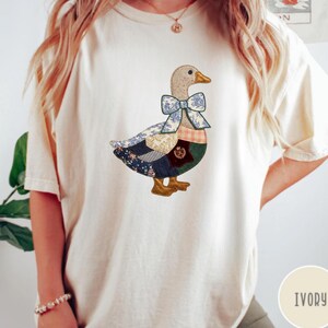 May include: Ivory-colored t-shirt featuring a patchwork duck design. The duck has a beige head, a blue bow, and a body composed of various fabric patterns. The shirt has short sleeves and a relaxed fit.