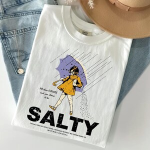 May include: White t-shirt with a graphic of a girl under a purple umbrella, with the word "SALTY" in large black letters. The shirt also has the text "All these flavors and you choose to be" and "This salt does not supply iodine..."