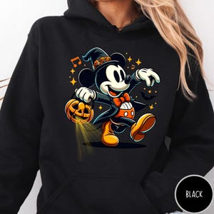 May include: A black hooded sweatshirt featuring a graphic of a cartoon mouse wearing a witch hat with small skull accents, an orange bow tie, and orange shoes. The mouse holds a glowing jack-o'-lantern, surrounded by yellow stars and music notes.