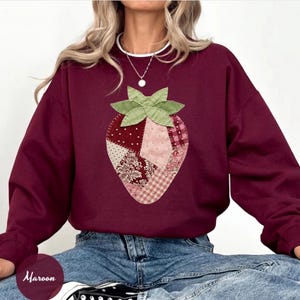 May include: Maroon sweatshirt featuring a patchwork strawberry design. The strawberry is composed of various fabric patterns in shades of red, pink, and white, with green leaves at the top. The word "Maroon" is written in cursive at the bottom.