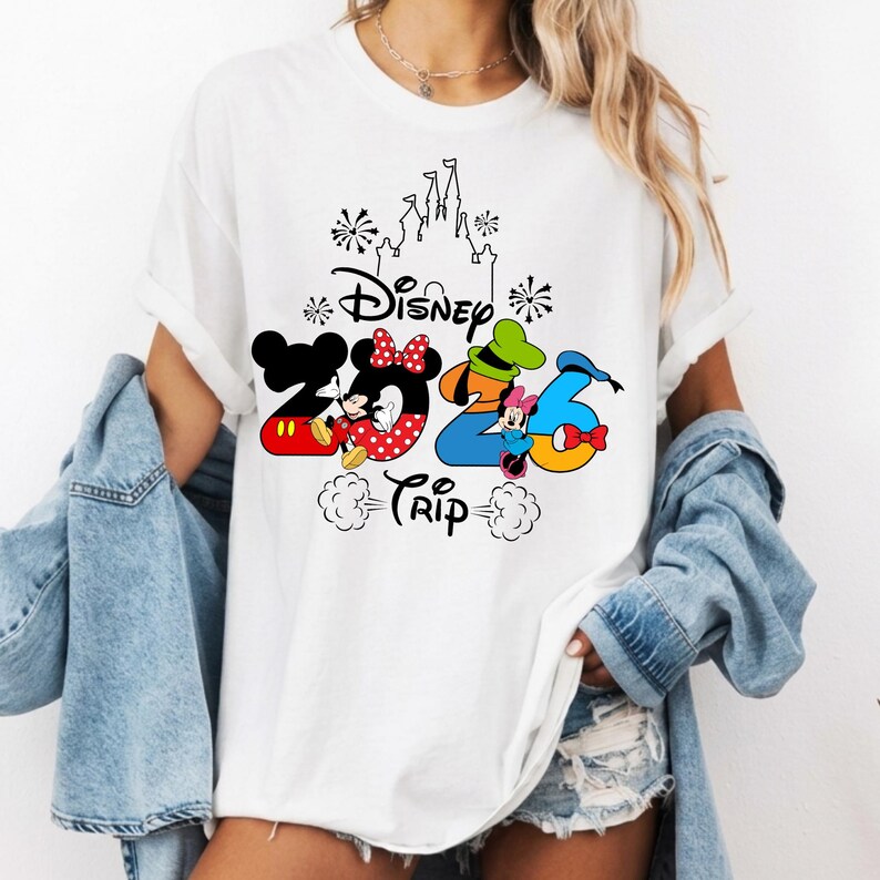 May include: A white short-sleeve t-shirt featuring a graphic design with the text Disney 2026 Trip. The design includes a black outline of a castle, fireworks, and the numbers 2026 filled with colourful illustrations of Mickey and Minnie Mouse.