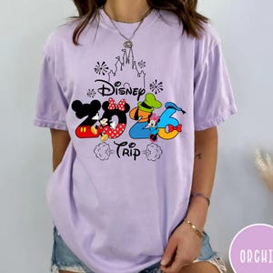 May include: A light purple short-sleeve t-shirt featuring a graphic print of a castle outline, fireworks, and the text Disney 2026 Trip. The design includes colourful illustrations of Mickey Mouse, Minnie Mouse, Goofy, and Donald Duck characters.
