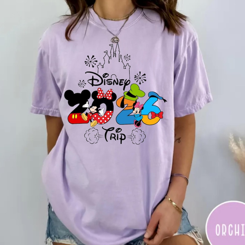 May include: A light purple short-sleeve t-shirt featuring a graphic print of a castle outline, fireworks, and the text Disney 2026 Trip. The design includes colourful illustrations of Mickey Mouse, Minnie Mouse, Goofy, and Donald Duck characters.