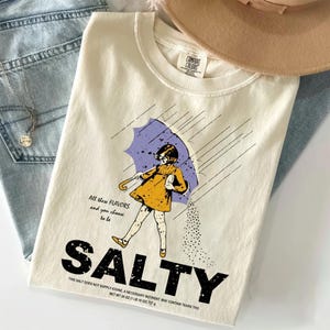 May include: Cream-colored t-shirt with a graphic of a girl under a purple umbrella, with the word "SALTY" in large black letters. The shirt also has the text "All these flavors and you choose to be".