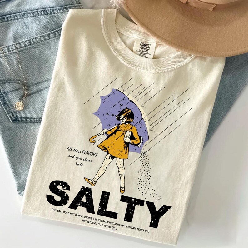 May include: Cream-colored t-shirt with a graphic of a girl under a purple umbrella, with the word "SALTY" in large black letters. The shirt also has the text "All these flavors and you choose to be".